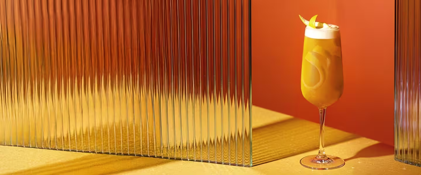A glass is being filled with a foamy drink beside a bottle of Johnnie Walker Blue Label whisky, a glass of whisky, a vase with yellow flowers, fruit, and snacks on a white tablecloth.