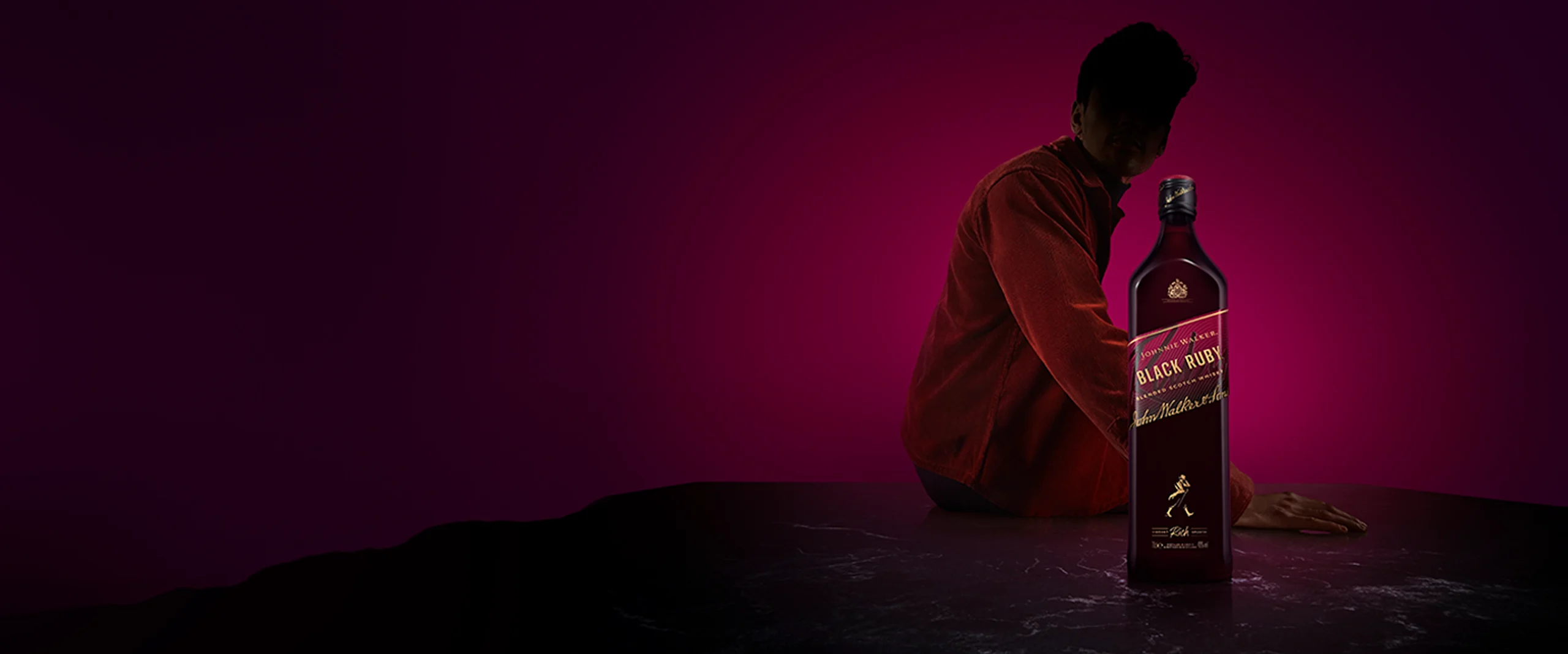 A dimly lit image features a dark bottle with a gold label reading Black Label on a marbled surface. In the background, a person in a red shirt is partially visible against a purple background.