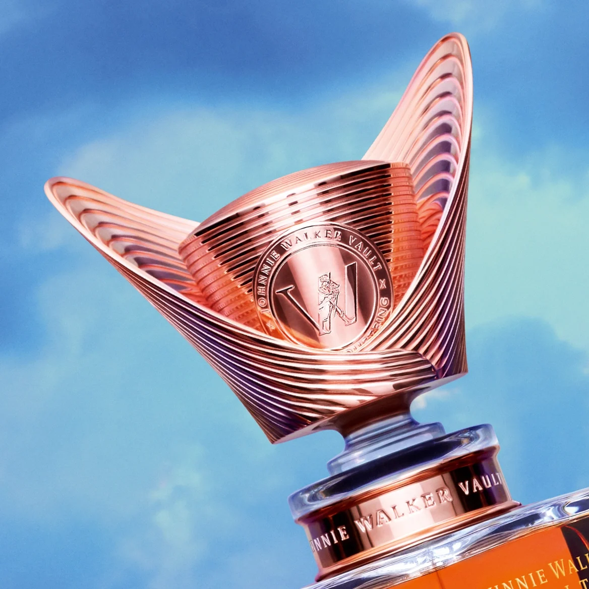 A luxurious Johnnie Walker whisky bottle with a metallic, sculpted design sits on a reflective surface against a dramatic, colorful sky background.
