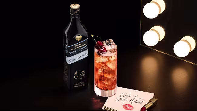 A bottle of Johnnie Walker Black Label whisky and a tall cocktail garnished with a cherry are placed on a silver tray atop a reflective black surface, with a black leather sofa in the background.