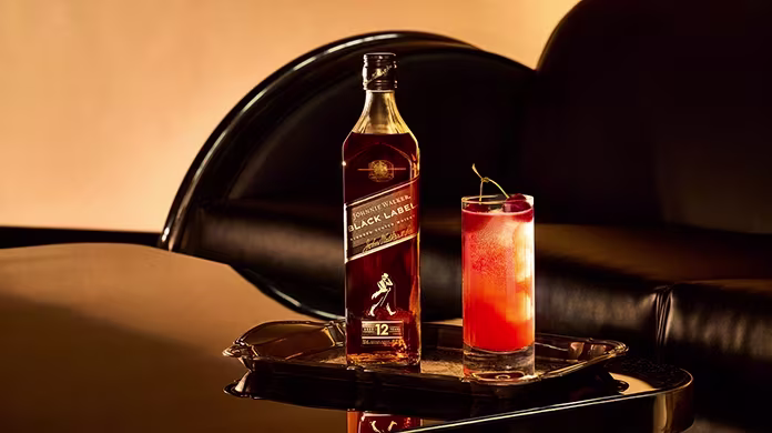 Uma garrafa de uísque Johnnie Walker Black Label e um coquetel alto, decorado com uma cereja, são colocados em uma bandeja de prata sobre uma superfície preta refletora, com um sofá de couro preto ao fundo.