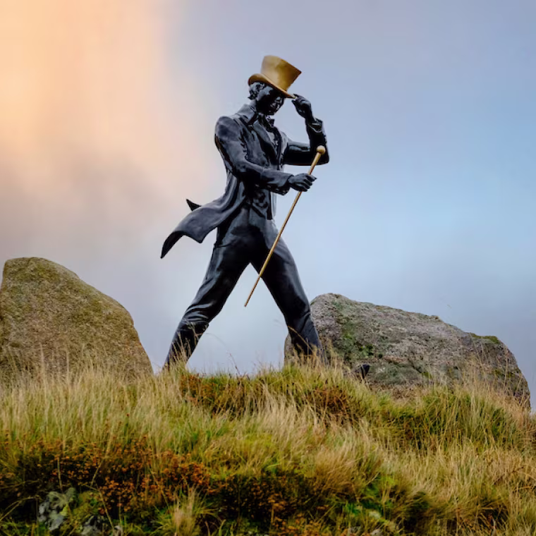A statue of a man in a suit and top hat holding a cane stands on grassy terrain between large rocks, set against a cloudy sky at sunset.