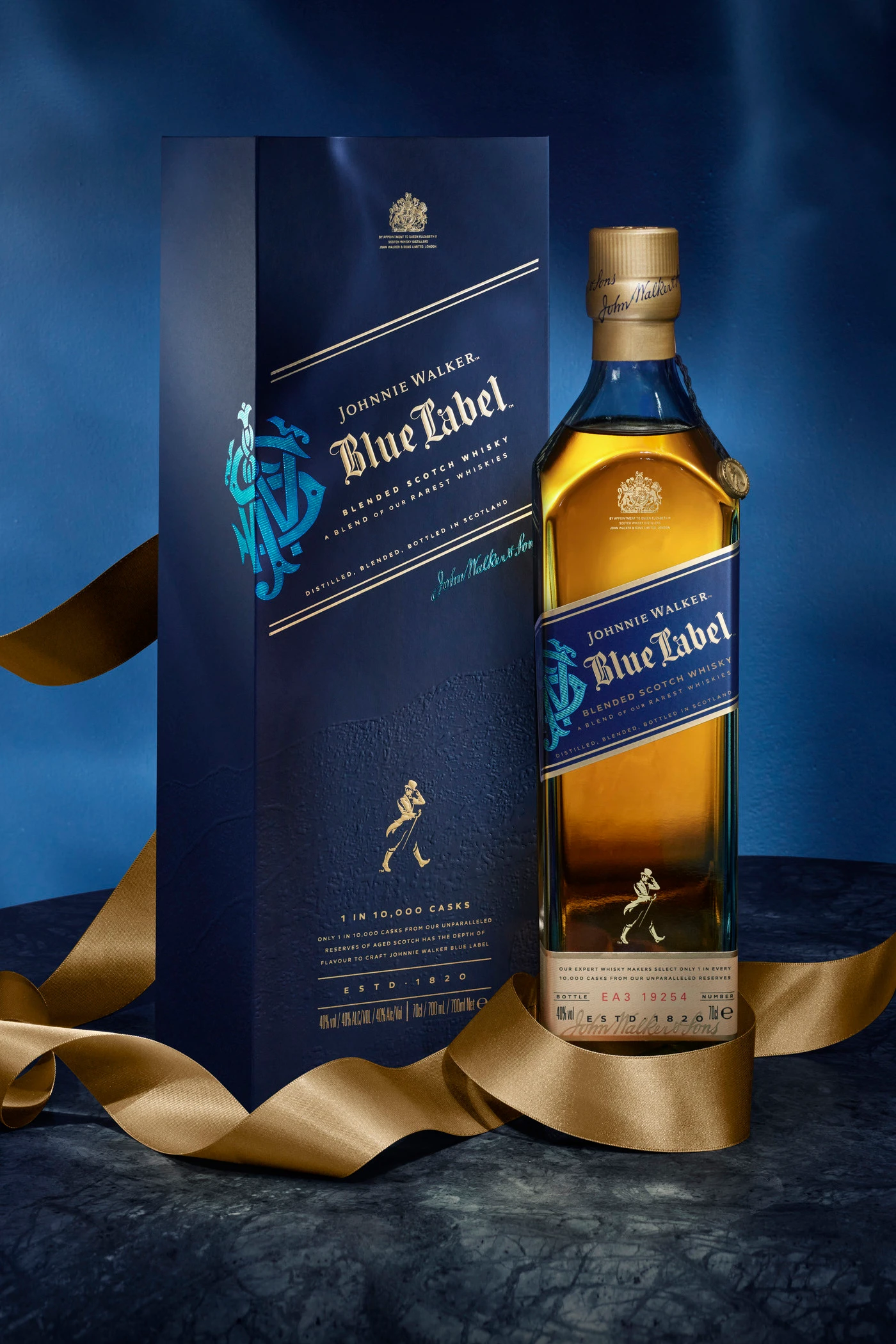 Una botella de whisky Johnnie Walker Blue Label se exhibe junto a su caja azul, con una cinta dorada elegantemente alrededor sobre una superficie oscura y texturizada.