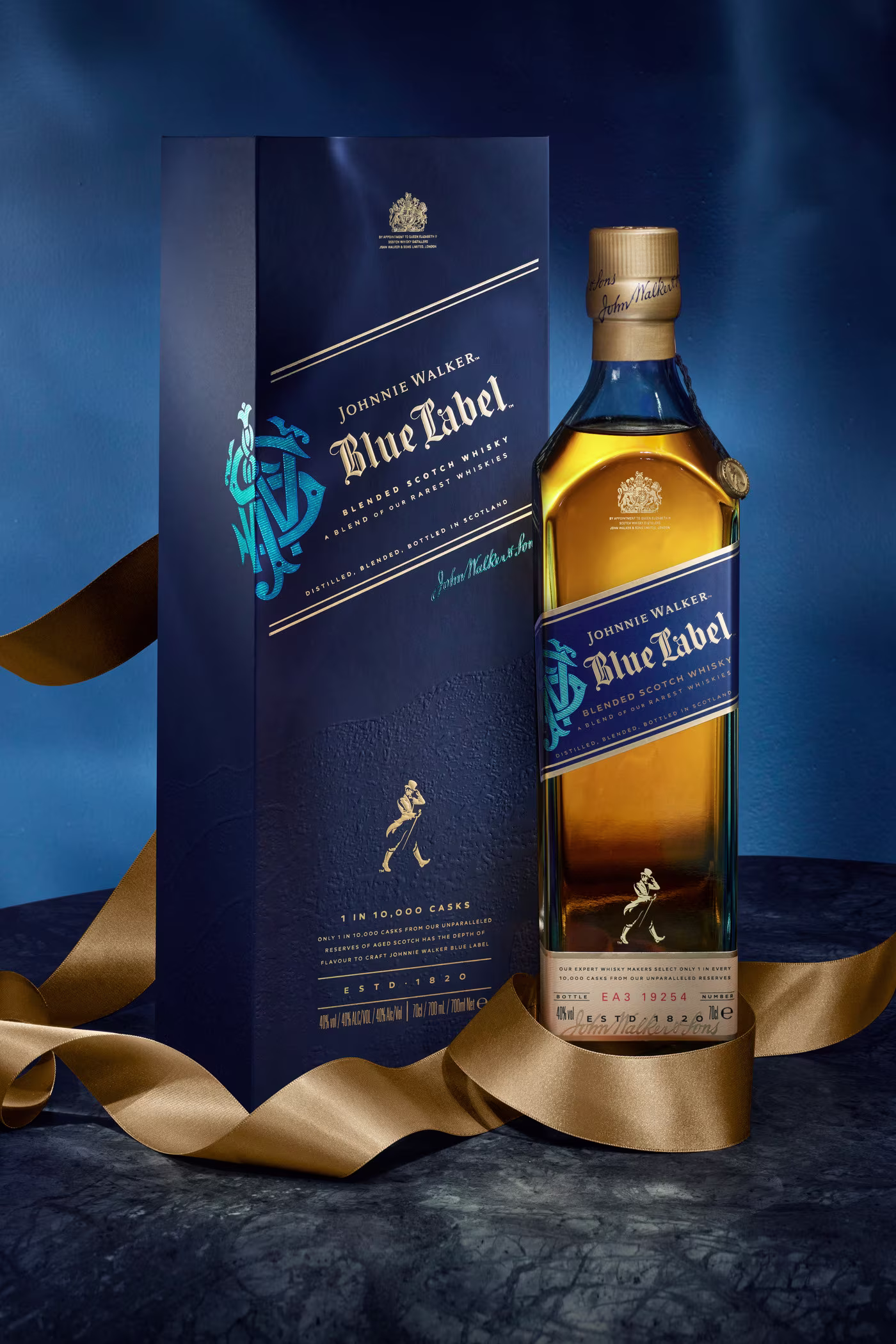 Изложена е бутилка уиски Johnnie Walker Blue Label с елегантна тъмносиня опаковка. Златна панделка се увива артистично около бутилката и кутията, поставена на наситен син фон.