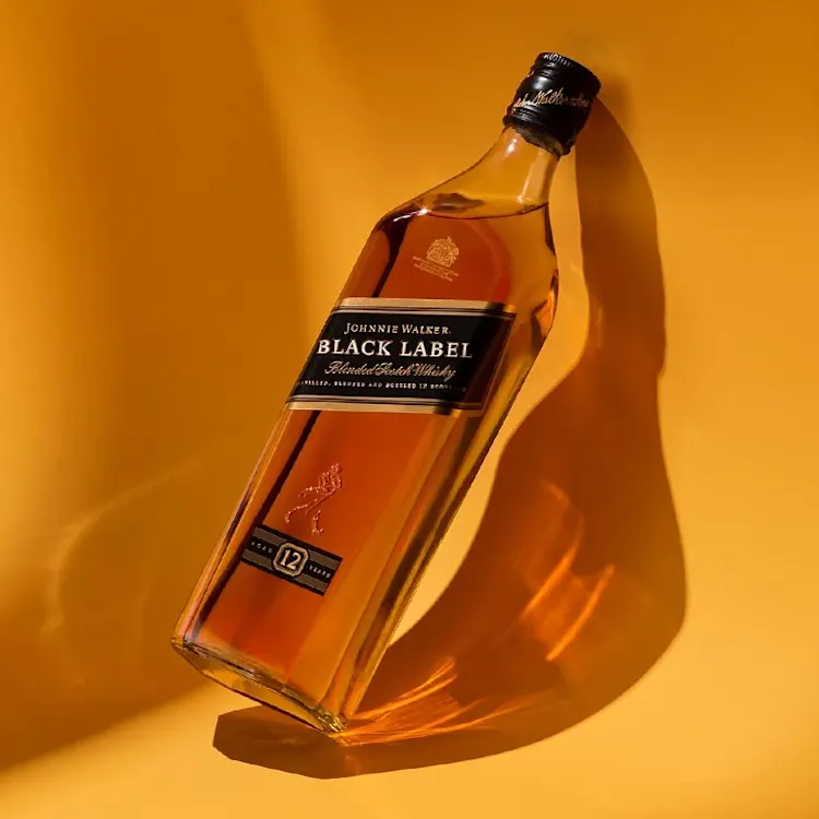 Uma garrafa de uísque Johnnie Walker Black Label é colocada sobre uma superfície amarela, com uma sombra projetada ao lado. O uísque tem uma cor âmbar rica e o rótulo indica que é um uísque escocês blended de 12 anos.