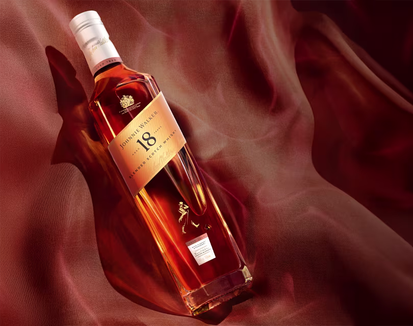 A bottle of Johnnie Walker 18-year-old blended Scotch whisky lies on a silky, flowing brown-red fabric background, with warm lighting highlighting the amber color of the liquid.