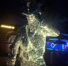 Una estatua de mosaico reflectante de un hombre con sombrero y traje, iluminada de noche. El fondo es oscuro, con luces de neón y letreros parcialmente visibles.