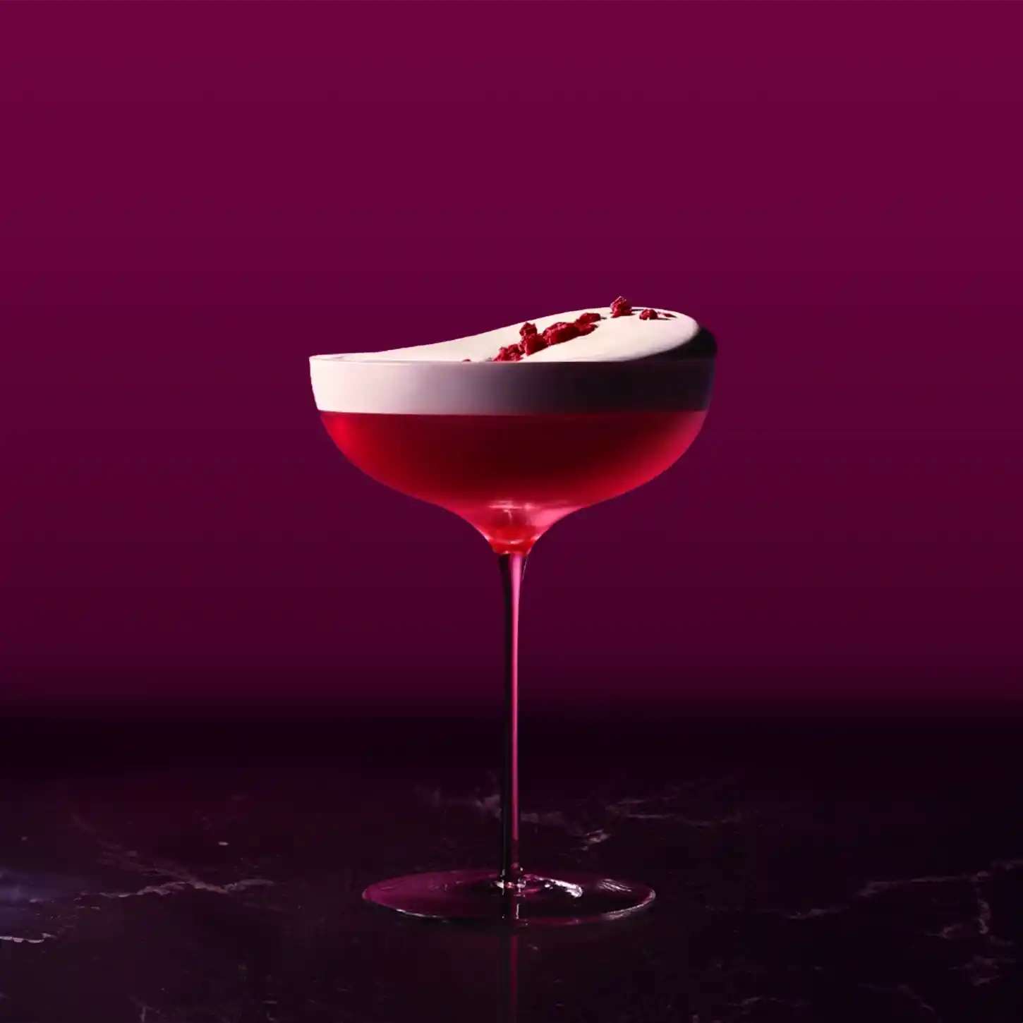 A vibrant cocktail in a coup glass against a dark purple background. The drink is dark red with a frothy white top, garnished with small red bits. The thin stem and wide bowl create an elegant presentation.