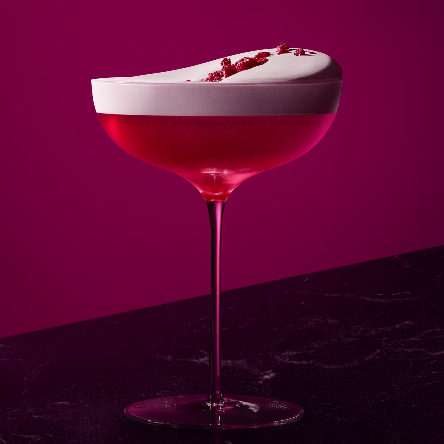 Ein lebendiger Cocktail mit einem satten Rosaton, bedeckt von dickem weißen Schaum und garniert mit kleinen roten Fruchtstücken. Das Getränk befindet sich in einem eleganten Glas vor einem einfarbigen magentafarbenen Hintergrund auf einer dunklen Fläche.