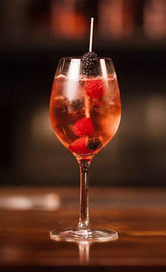 Ein Weinglas, gefüllt mit einem rosafarbenen Cocktail, Eiswürfeln, Himbeeren und Brombeeren, garniert mit einem Spieß, der eine Brombeere hält, auf einer Holzoberfläche vor verschwommenem Hintergrund.