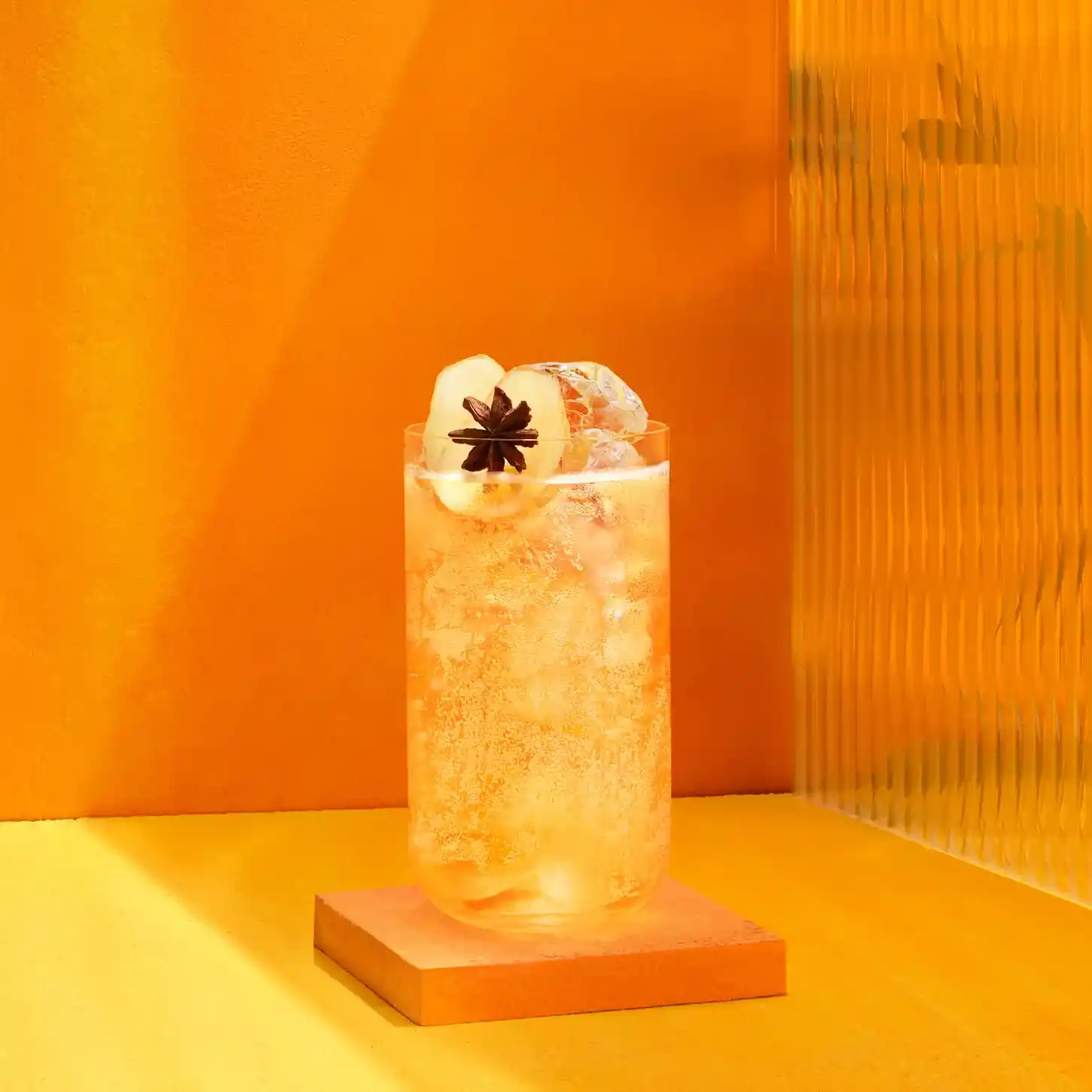 Un vaso alto lleno de una refrescante bebida cítrica, adornado con hielo, rodajas de cítricos y anís estrellado, se encuentra sobre un posavasos de color naranja. El fondo es de un naranja vibrante, lo que crea una atmósfera luminosa y soleada.