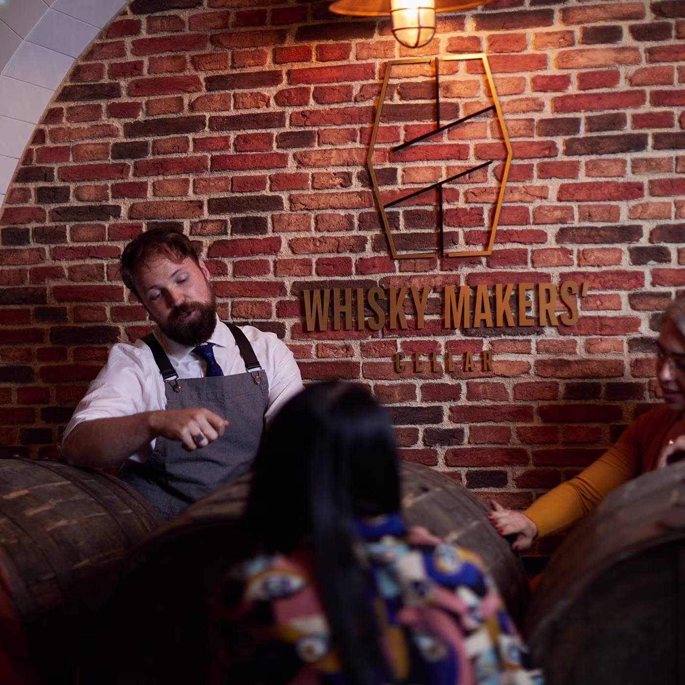 A man in an apron talks to people by whisky barrels under a Whisky Makers Cellar sign on a brick wall.