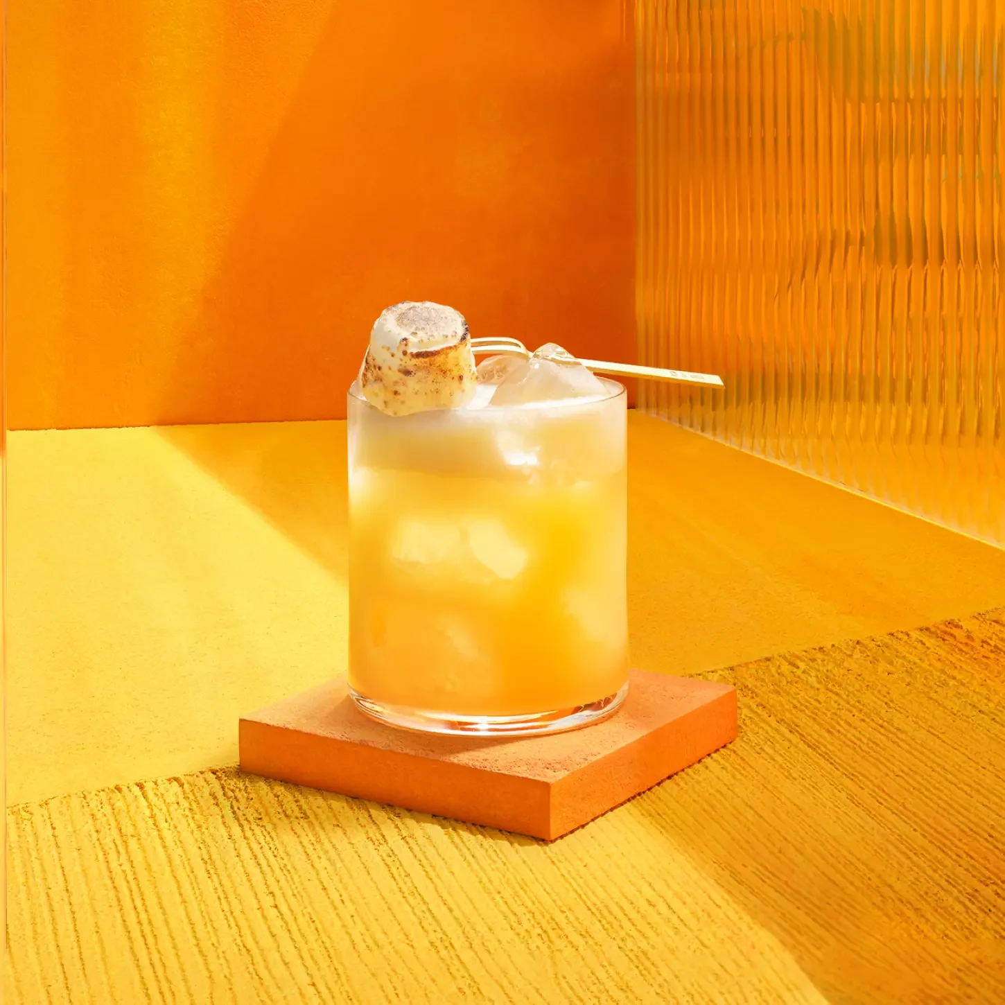 Un verre de cocktail glacé orange-jaune est agrémenté d'une guimauve grillée sur une brochette. La boisson est présentée sur un dessous de verre orange, sur un fond orange et jaune vif et texturé.