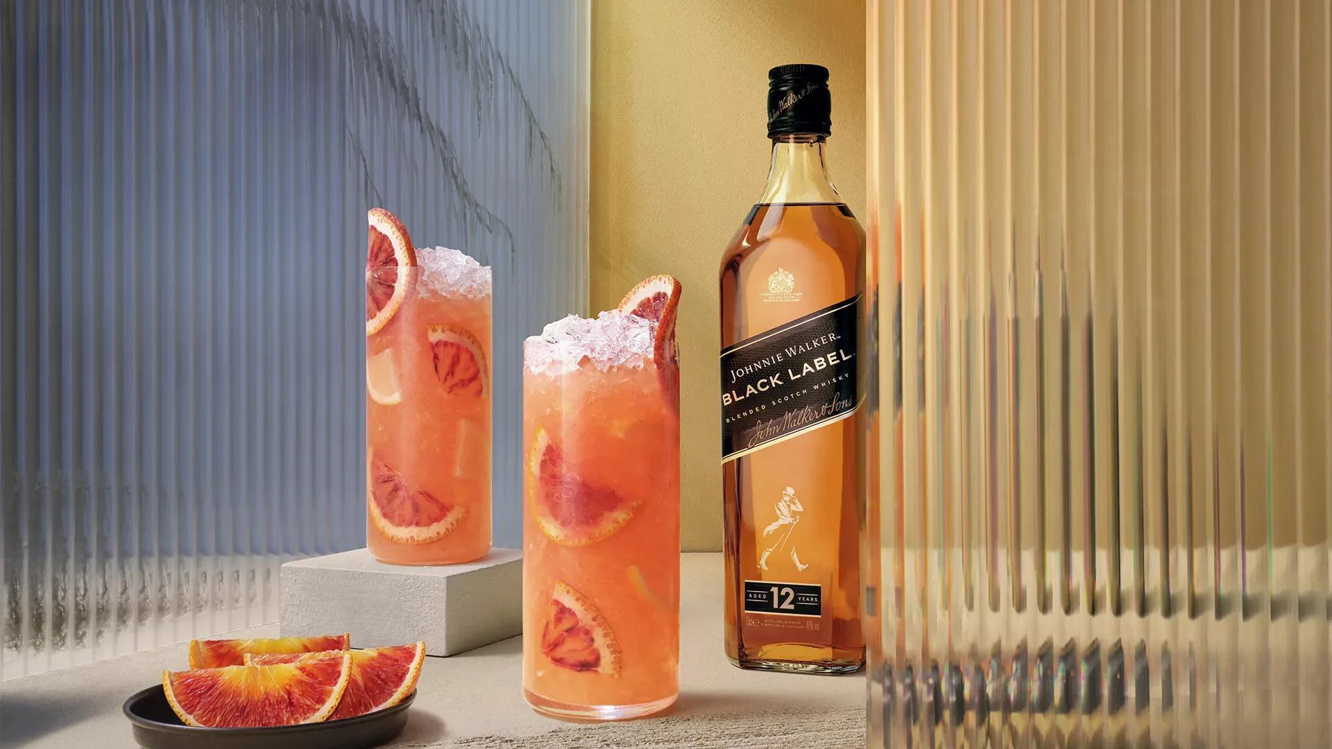 A bottle of Johnnie Walker Black Label whisky stands next to two tall glasses filled with an orange-red cocktail, ice, and blood orange slices. Nearby is a small plate with more blood orange wedges.