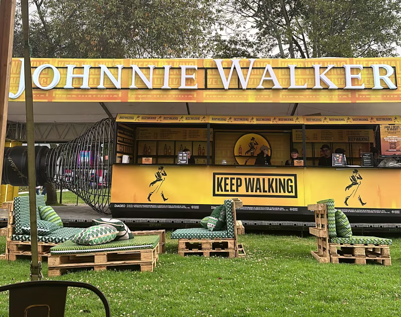 Un puesto de Johnnie Walker en un espacio público luce un gran cartel de "Keep Walking" y la marca en amarillo y negro. Se ve una barra con personal detrás, y entre los elementos decorativos se incluyen una gran chapa de botella y asientos estampados.