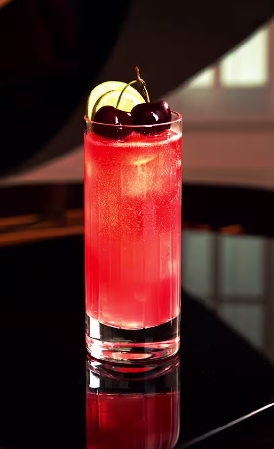 A tall glass of bright pink sparkling drink garnished with fresh cherries and lime slices, set on a glossy black surface with soft natural light in the background.
