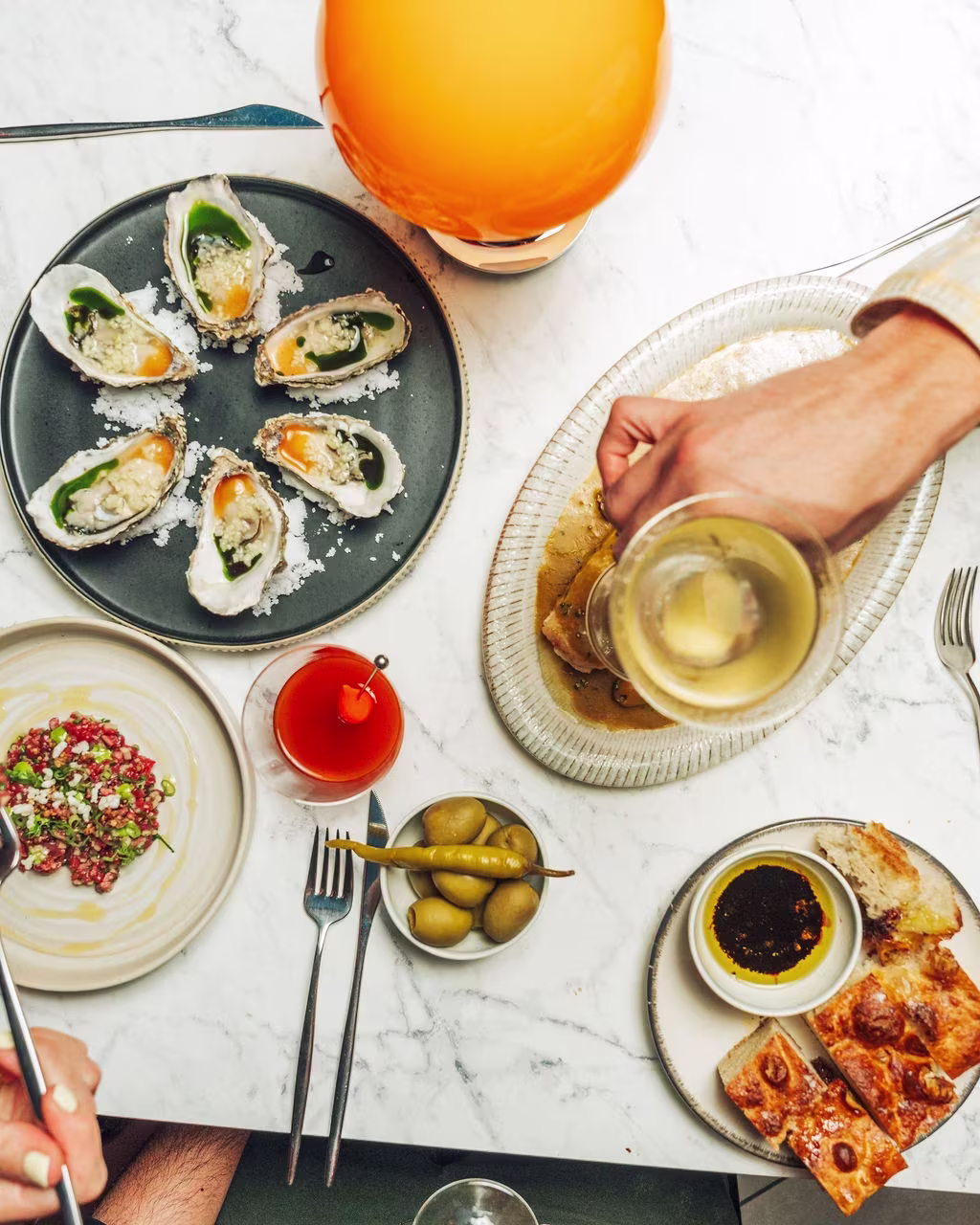 Top-down view of a table with oysters, bread, olives, a cocktail, wine being poured, and tartare.