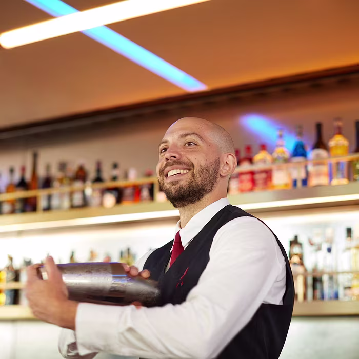 Smiling bartender shaking a cocktail shaker behind a bar with bottles on shelves in the background.