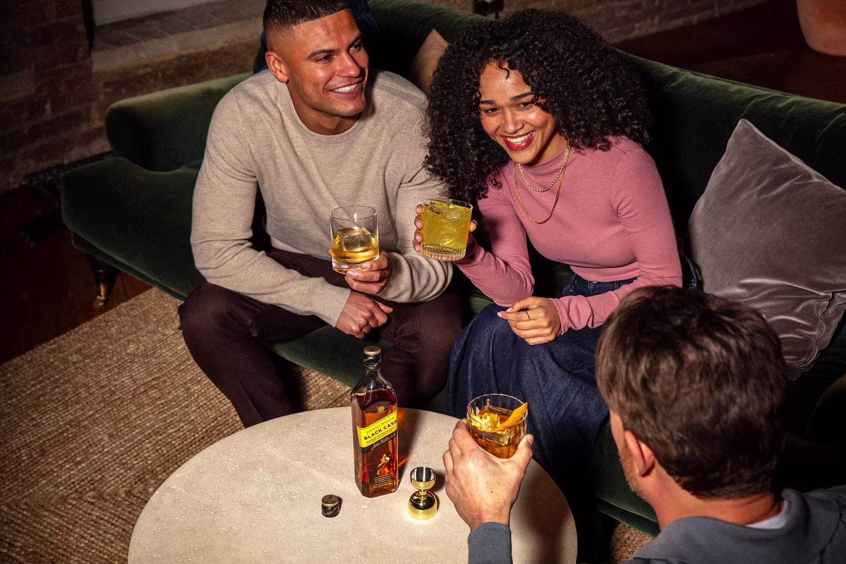 Three people sit on a green couch and around a small table, smiling and holding glasses of whiskey. A bottle of Johnnie Walker and a jigger rest on the table in front of them.