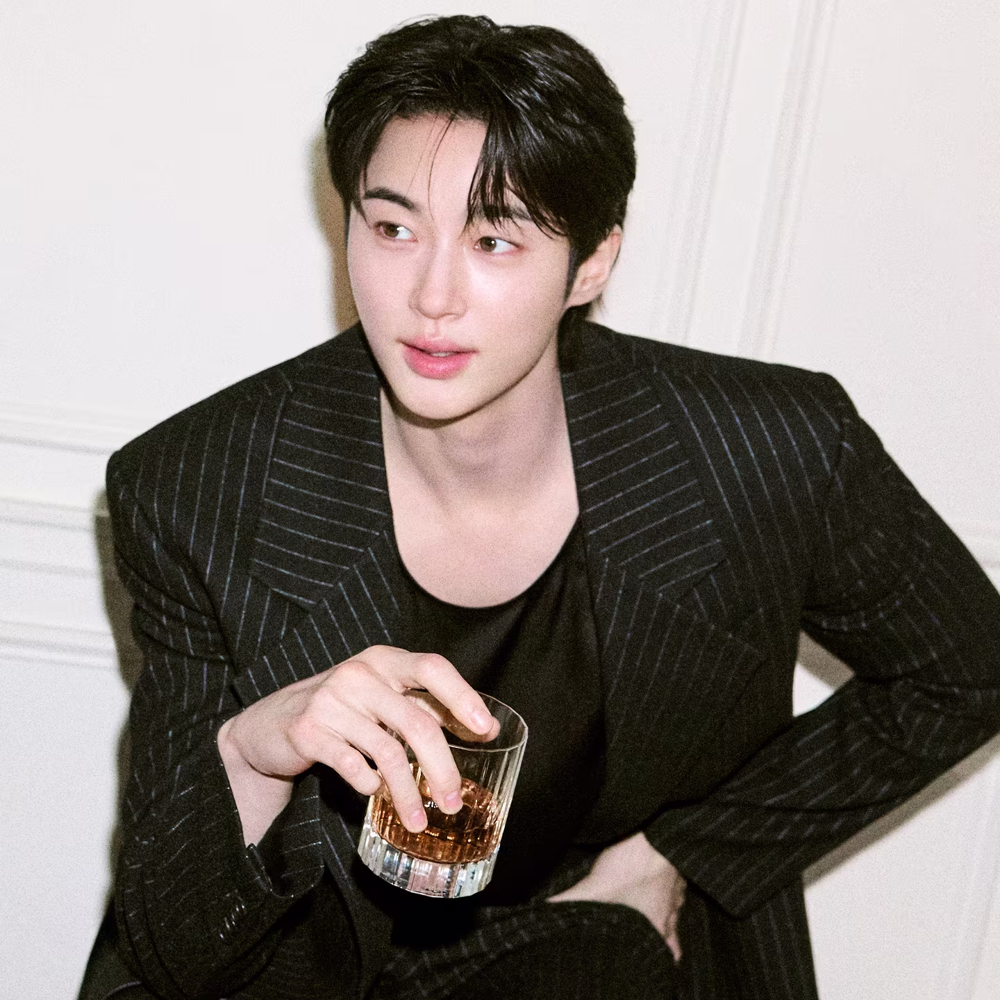 A person in a pinstripe suit holds a glass of brown liquid while sitting against a white wall.