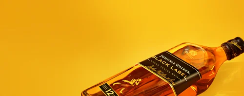 A bottle of Johnnie Walker Black Label Scotch whiskey lies on its side against a bright yellow background. The label is black with gold text, featuring the iconic running man logo. The cap of the bottle is black with a gold accent.