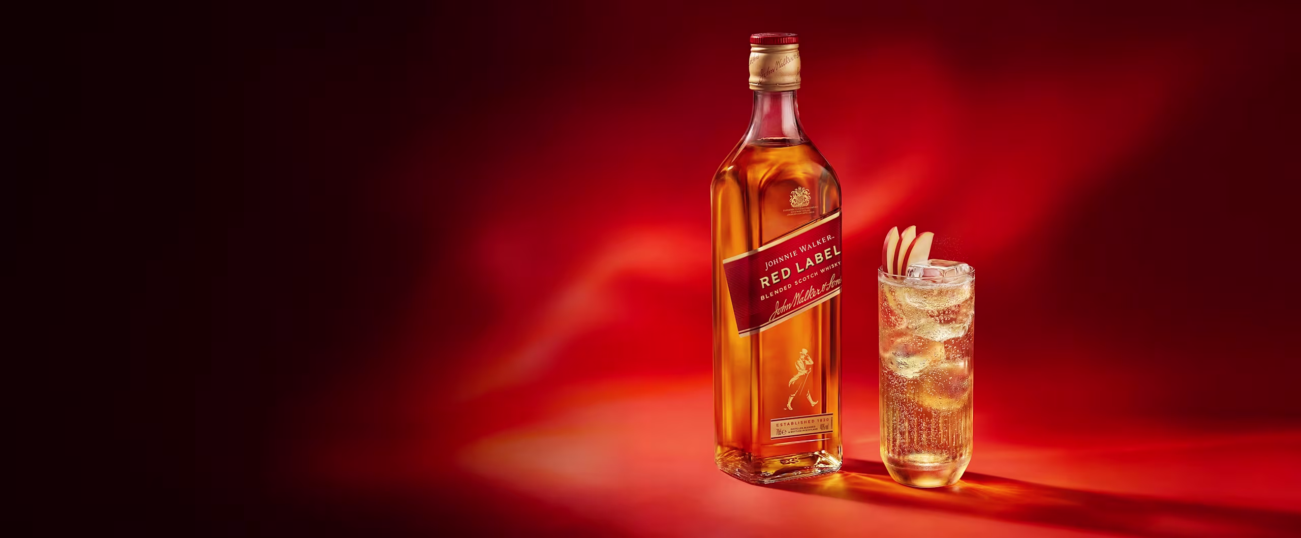 Una botella de whisky Johnnie Walker Red Label junto a un vaso de whisky con hielo y una guarnición, sobre un fondo rojo.