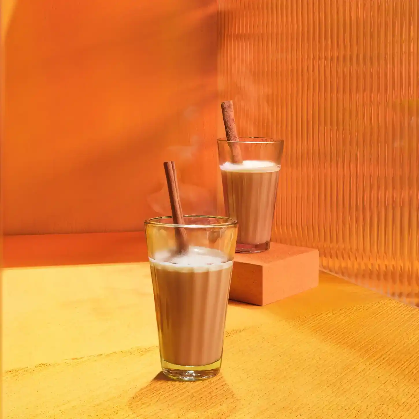 Dos vasos de leche caliente especiada con palitos de canela sobre una superficie amarilla.