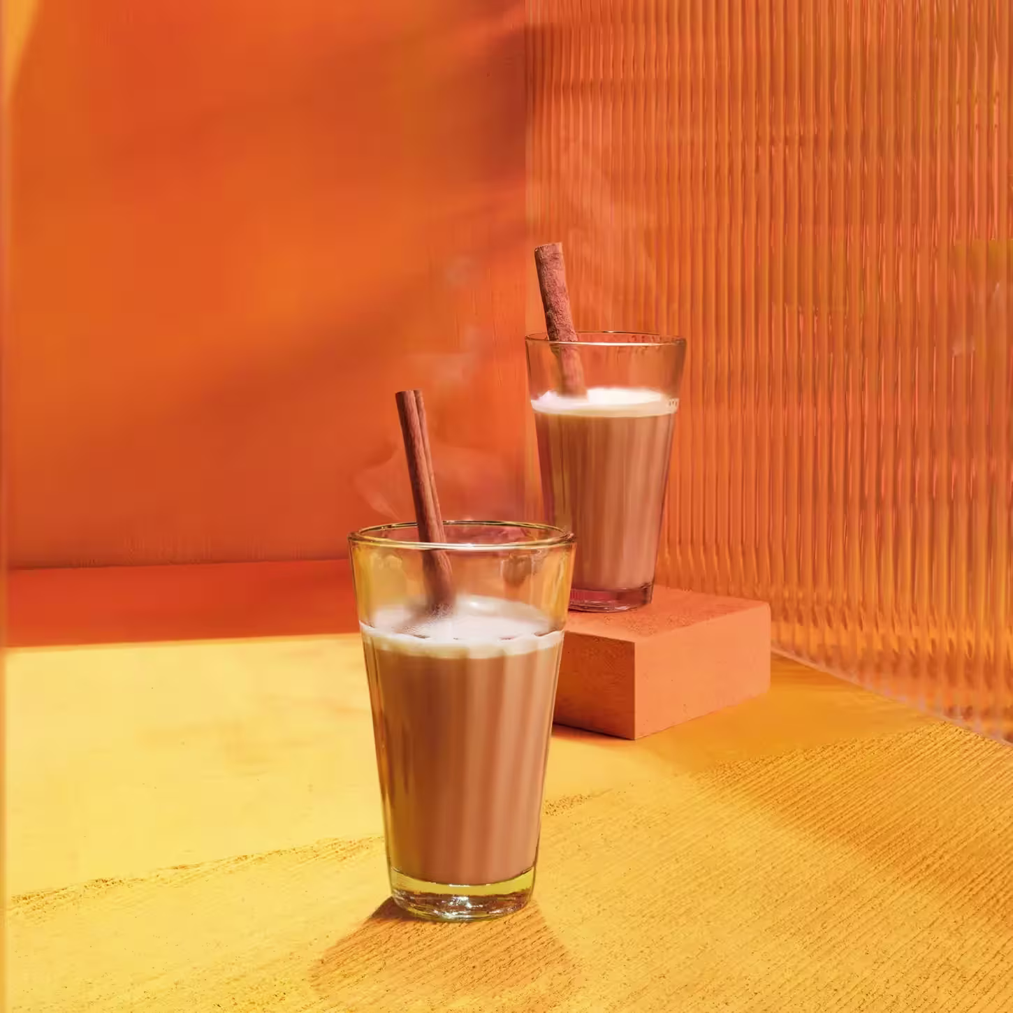 Deux verres de chai latte fumants sont posés sur une surface orange. Chaque verre est décoré d'un bâton de cannelle. Le fond, orné d'un mur texturé orange, crée une atmosphère chaleureuse.