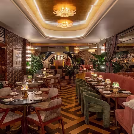 Elegant restaurant interior with plush seating, patterned wood floors, round tables set with plates and decorative lamps, plants, warm lighting, and gold-accented ceiling, creating a luxurious and inviting atmosphere.