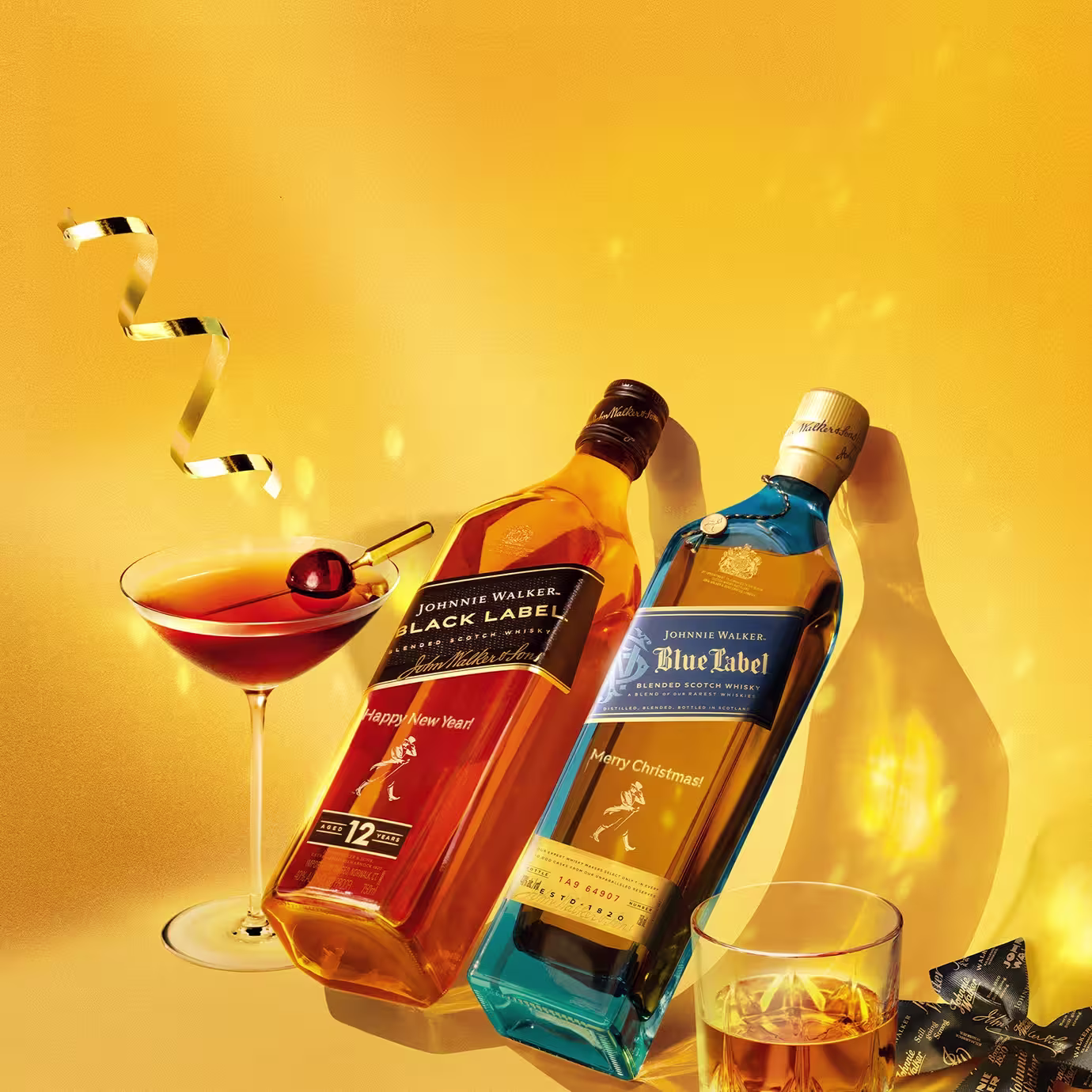 Two bottles of Johnnie Walker whisky, Black Label and Blue Label, are arranged on a golden surface with a cocktail, a glass of whisky, a gold ribbon, and a blue gift bow, creating a festive holiday scene.