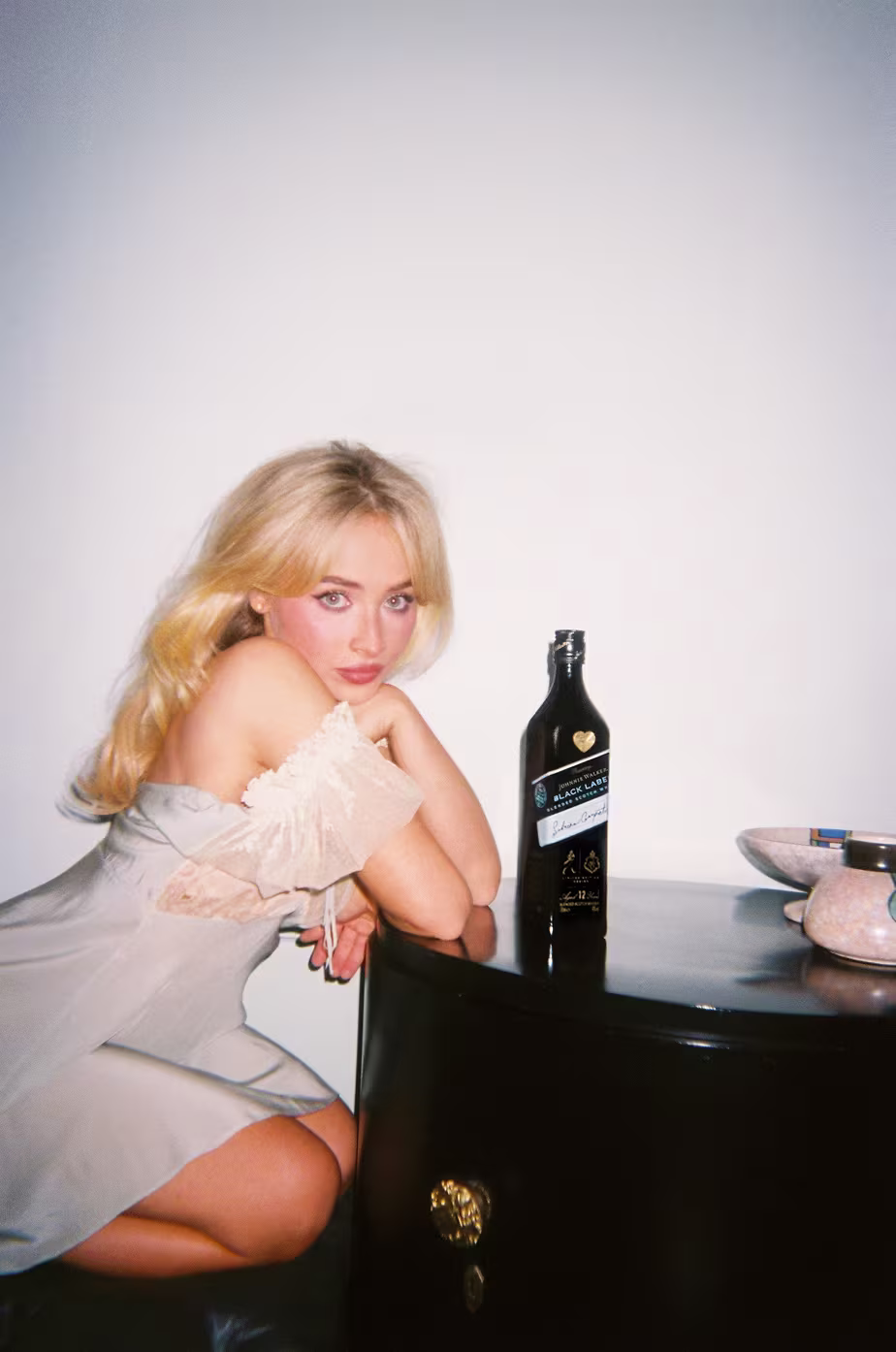 Sabrina Carpenter with long blonde hair in a light dress sits by a table with a dark bottle and a bowl.