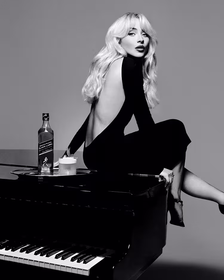 A woman in a backless black dress sits on a grand piano beside a bottle of whisky and a glass. The image is in black and white, with a stylish and elegant atmosphere.