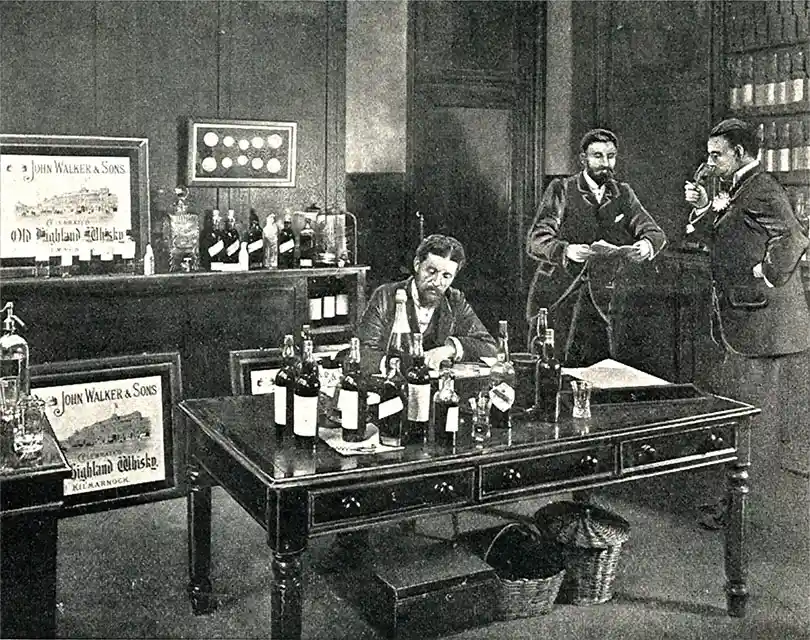 Uma foto antiga de três homens em uma sala cheia de garrafas e anúncios do uísque John Walker & Sons. Um homem está escrevendo em uma mesa enquanto os outros estão por perto. A sala tem painéis de madeira escura e quadros emoldurados nas paredes.