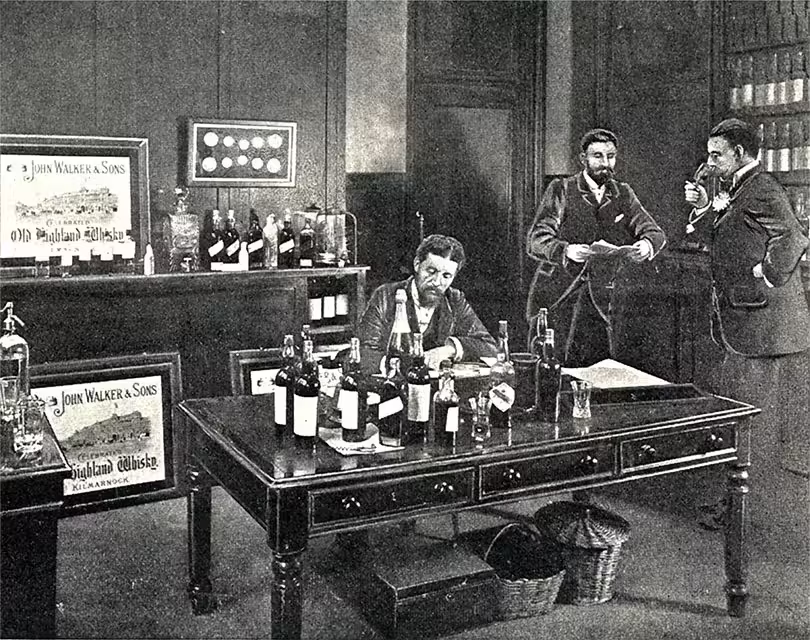 Black and white photo of three men in the office. A man is writing in front of a table covered with bottles. Two other men stood, one holding a phone. Posters and bottles are displayed on shelves in the background.