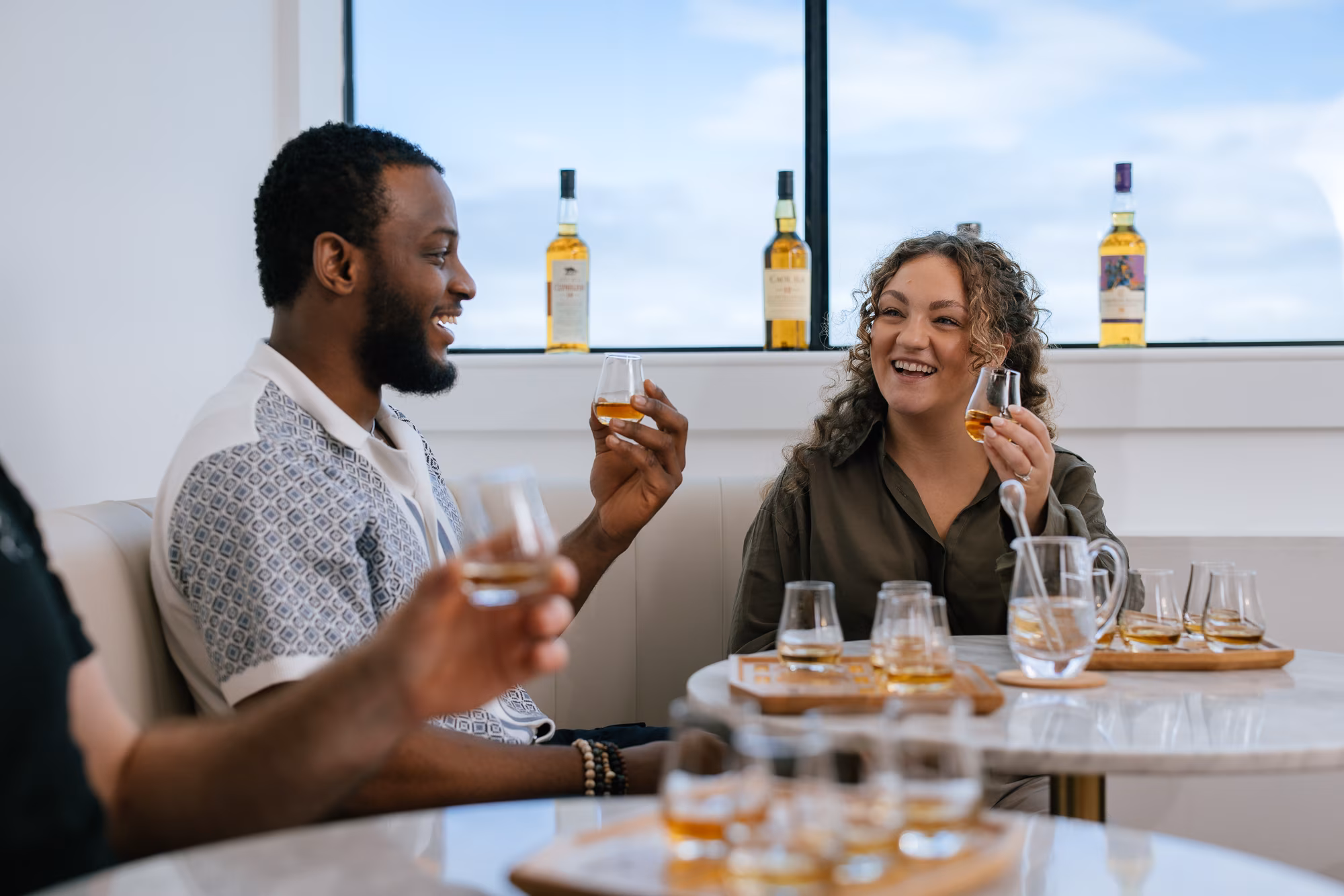 Two people smiling and clinking whiskey glasses together at a tasting, bottles and glasses on tables.