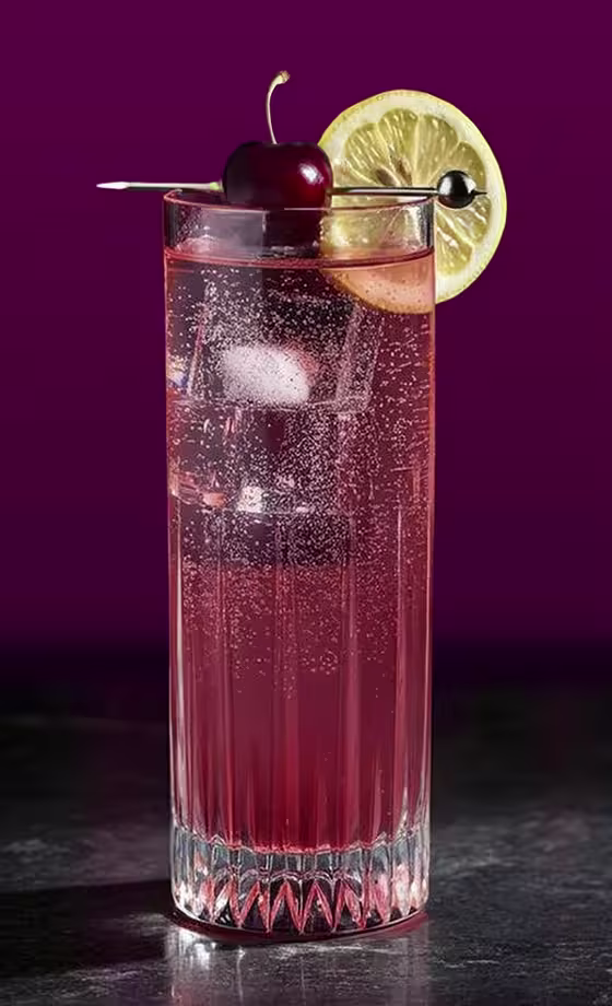 Un vaso alto de una bebida gaseosa roja con hielo está adornado con una rodaja de limón y una cereza en un palillo de cóctel, sobre un fondo violeta oscuro sobre una superficie de mármol negro.