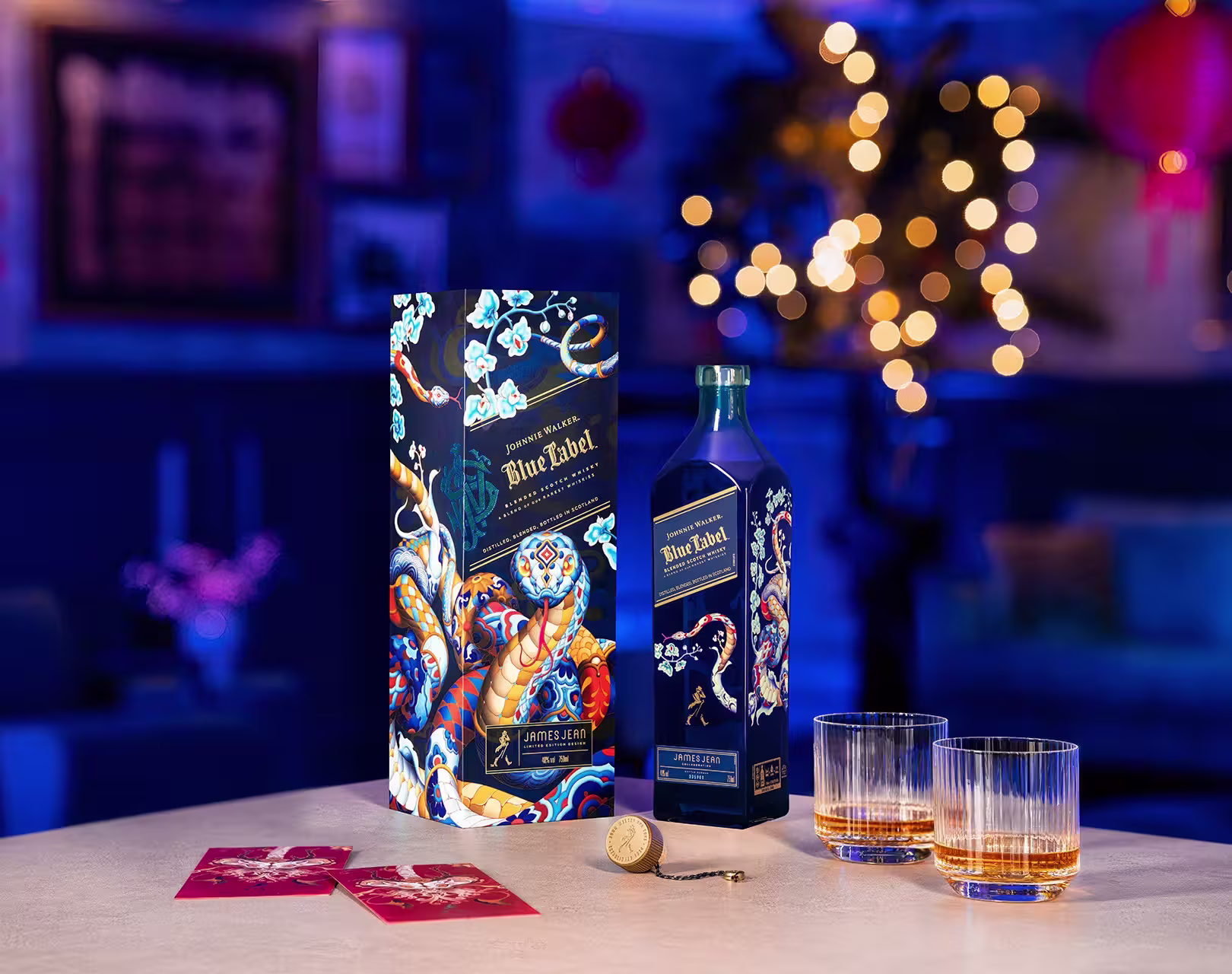 กล่องตกแต่งสวยงามและขวด Blue Label Year of the Snake Limited Edition วางอยู่บนโต๊ะพร้อมแก้ววิสกี้สองใบวางอยู่ข้างๆ พื้นหลังมีแสงไฟสลัวๆ พร้อมของตกแต่งวันหยุด เช่น ต้นไม้เรืองแสงและโคมไฟสีแดง