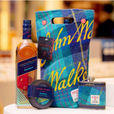 Una botella de whisky Johnnie Walker, una bolsa de transporte de tartán azul, una petaca, una lata y una etiqueta se exhiben sobre una mesa. La bolsa presenta un diseño a cuadros azules y amarillos con la palabra "Walker" escrita en amarillo.
