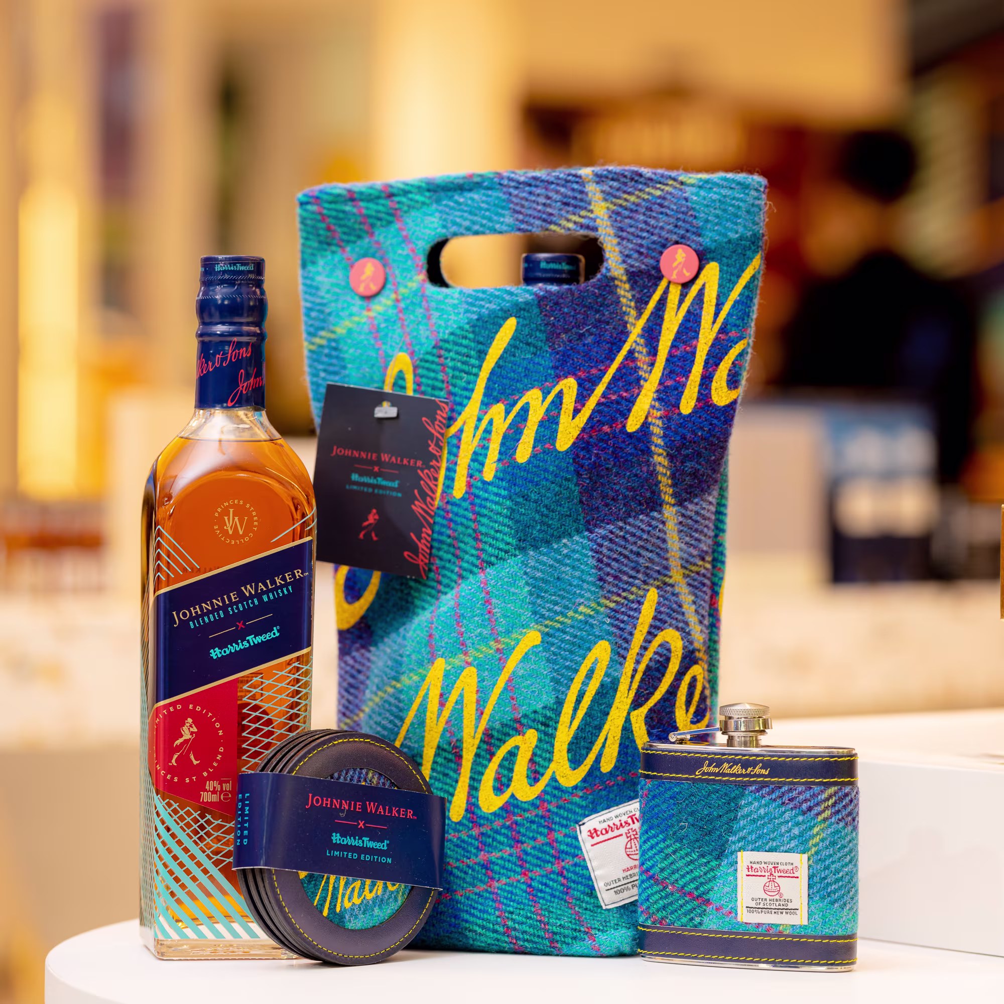 A bottle of Johnnie Walker whisky, a blue tartan carrying bag, a flask, a tin, and a label are displayed together on a table. The bag features a blue and yellow plaid design with “Walker” written in yellow.