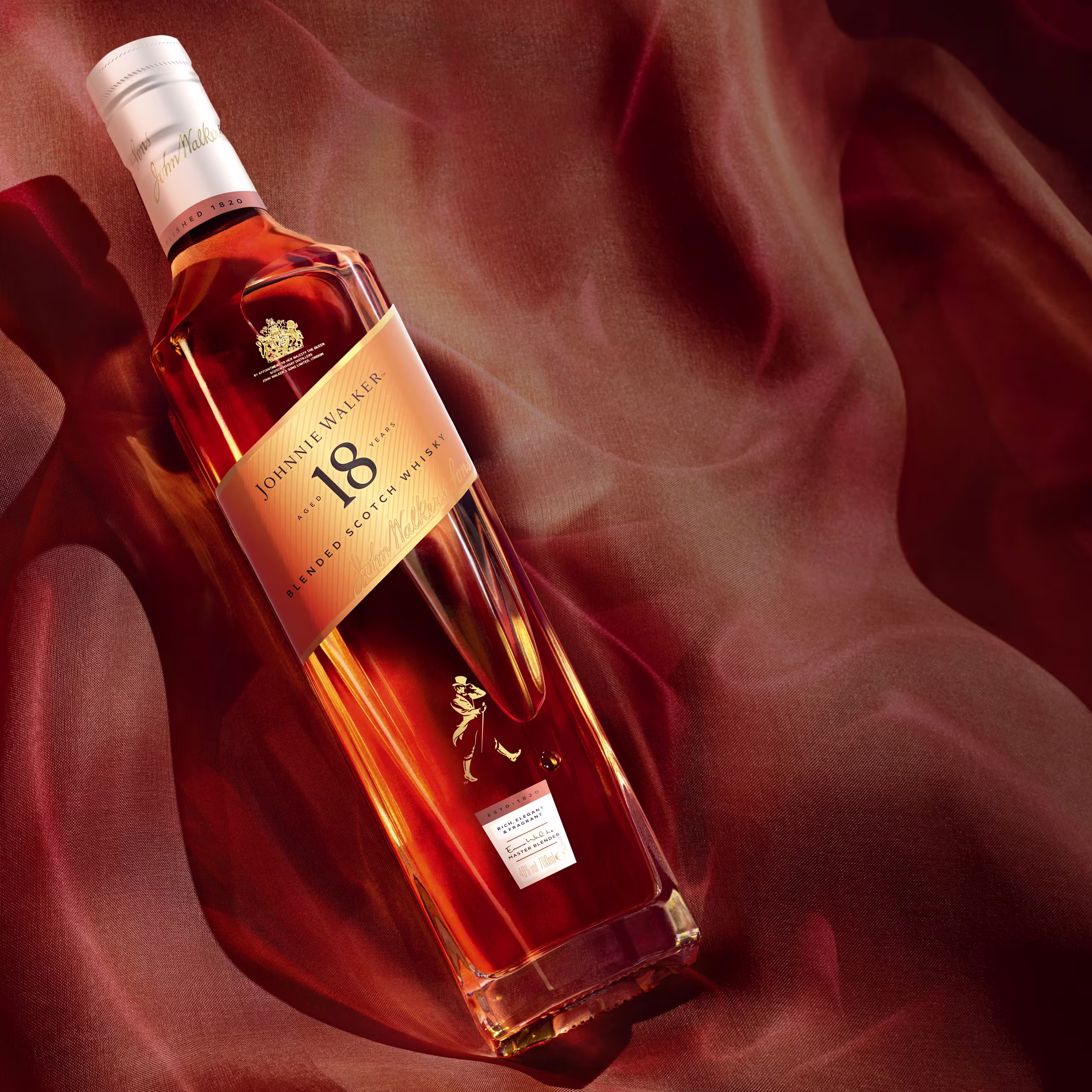 Une bouteille de whisky écossais mélangé Johnnie Walker de 18 ans repose sur un tissu rouge et brun fluide et soyeux, avec un éclairage chaleureux mettant en valeur le liquide ambré à l'intérieur de la bouteille.
