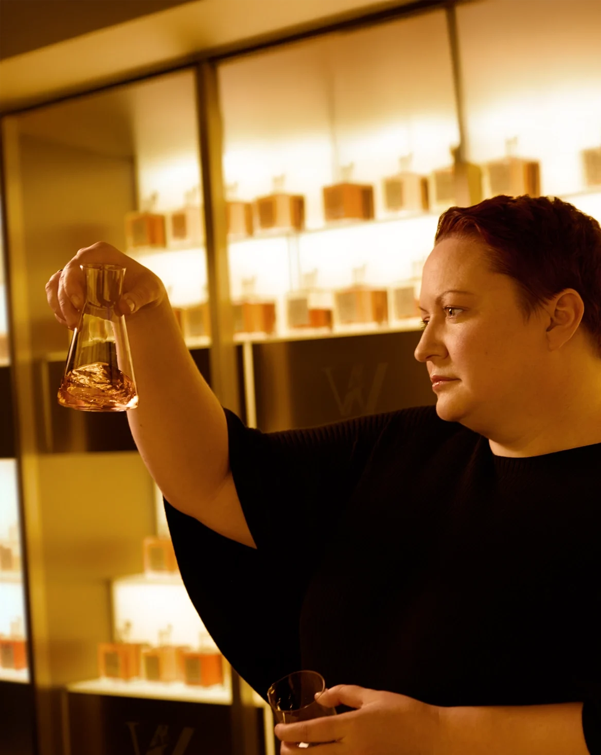 Dr. Emma Walker with short hair holds a glass vial, inspecting its contents, in a room filled with perfume bottles on shelves. The lighting is warm, creating an inviting ambiance. They are wearing a black outfit.