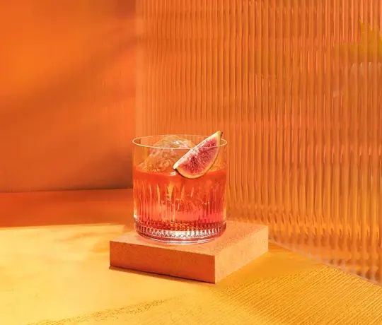 The Dark Toffee Boulevardier Cocktail. A glass of a vibrant orange cocktail with ice and a slice of fig on top, placed on a small wooden platform against a textured orange background. The setting is warm and minimalistic.