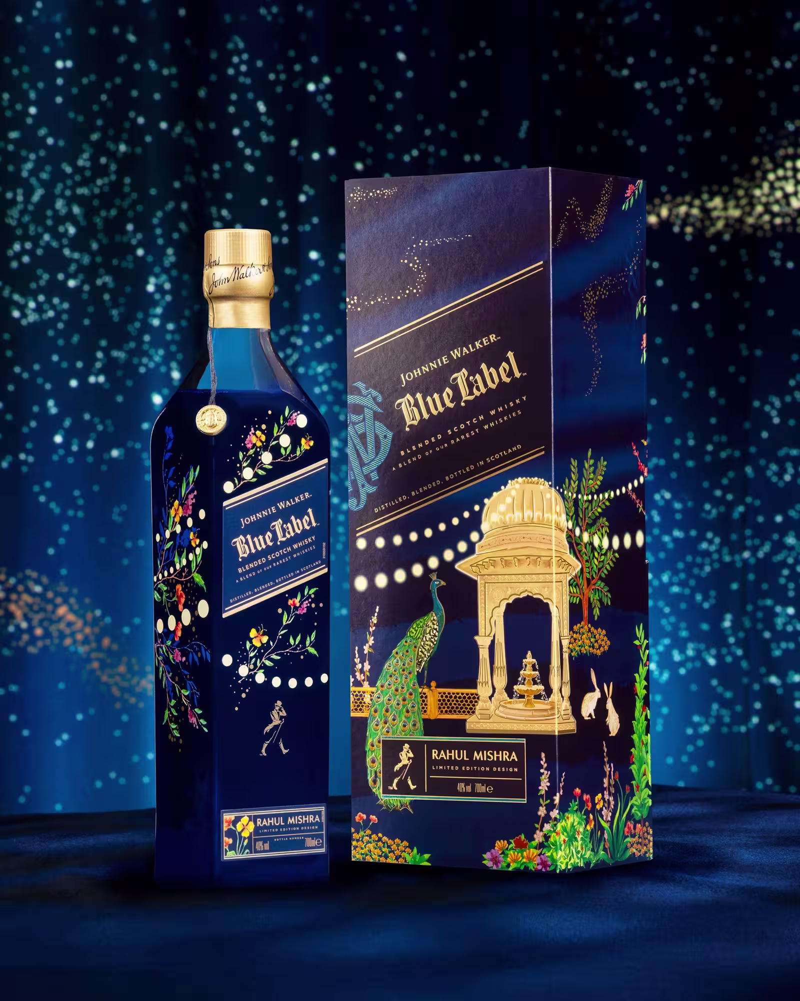 A blue bottle of Johnnie Walker Blue Label Limited Edition X Rahul Mishra stands beside its ornate box, both featuring intricate gold, floral, and animal designs against a starry blue backdrop. The box displays a peacock, gazebo, and elegant gold detailing