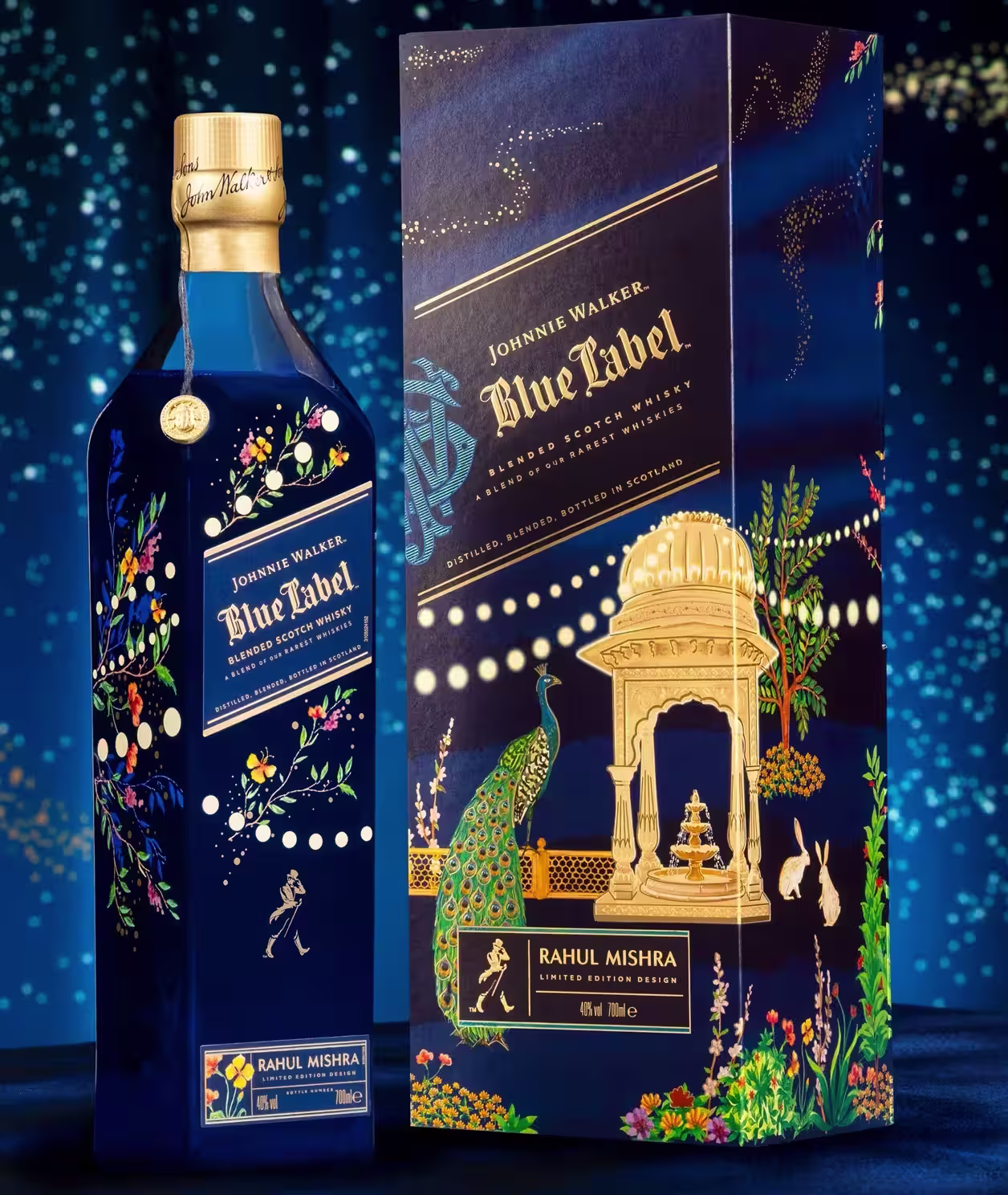 A blue bottle of Johnnie Walker Blue Label Limited Edition X Rahul Mishra stands beside its ornate box, both featuring intricate gold, floral, and animal designs against a starry blue backdrop. The box displays a peacock, gazebo, and elegant gold detailing