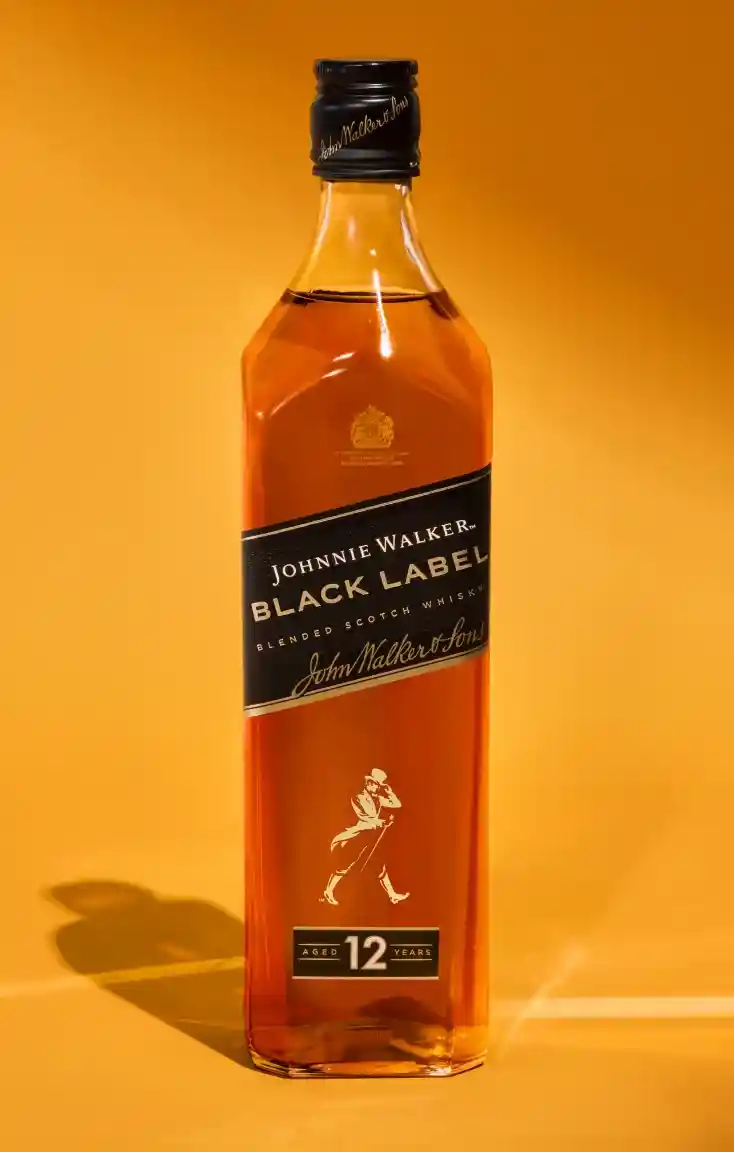 Uma garrafa de Johnnie Walker Black Label, um uísque escocês blended, colocada contra um fundo laranja quente. O rótulo exibe Johnnie Walker Black Label e 12 Years em dourado e preto, com o icônico logotipo do homem caminhando abaixo.