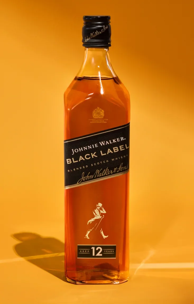 Бутилка блендирано шотландско уиски Johnnie Walker Black Label е изложена на топъл оранжев фон. На бутилката има черен етикет с логото на марката и надпис 12 години, указващ възрастта на уискито.