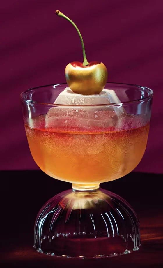 A cocktail in a glass with a large ice sphere and a cherry garnish sits on a table against a deep burgundy background with diagonal shadow lines.