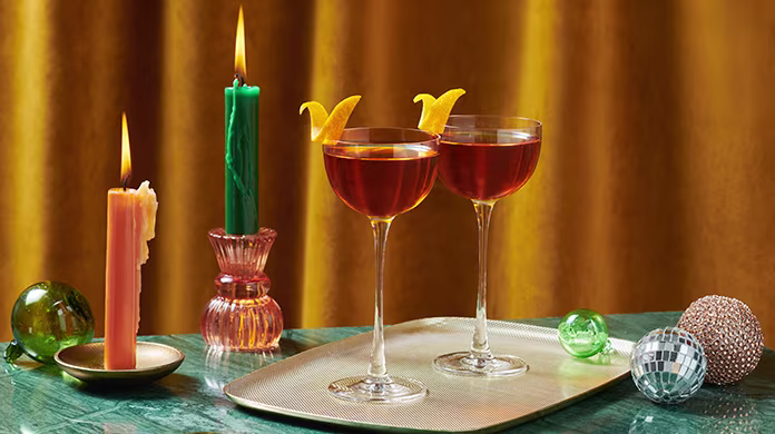 Dos cócteles en copas de cóctel con adornos de naranja reposan en una bandeja junto a velas de colores y decoraciones festivas, con un telón de fondo de cortinas doradas.