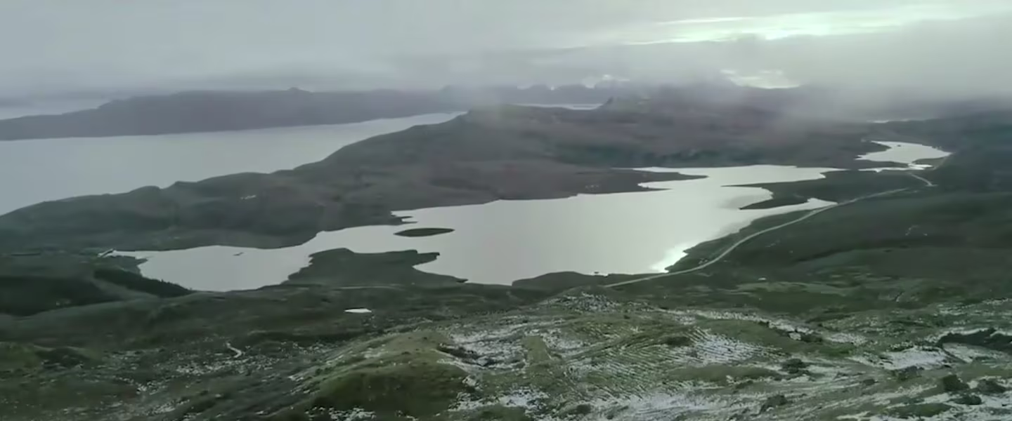 Vue panoramique d'un grand lac entouré de collines et de montagnes sous un ciel nuageux, avec quelques plaques de neige visibles au sol au premier plan. L'eau réfléchit la faible lumière du ciel.