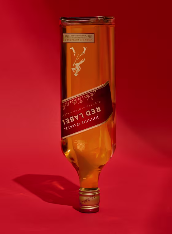 A bottle of Red Label whisky is positioned upside down against a vibrant red background, casting a shadow below. The label and branding are clearly visible, with the cap facing upwards.